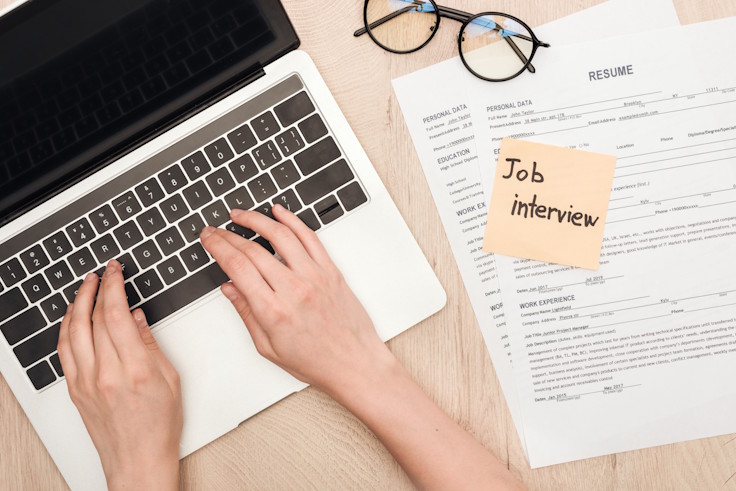 Hands typing on a laptop keyboard beside a resume, eyeglasses, and a sticky note with 'Job interview' written on it.
